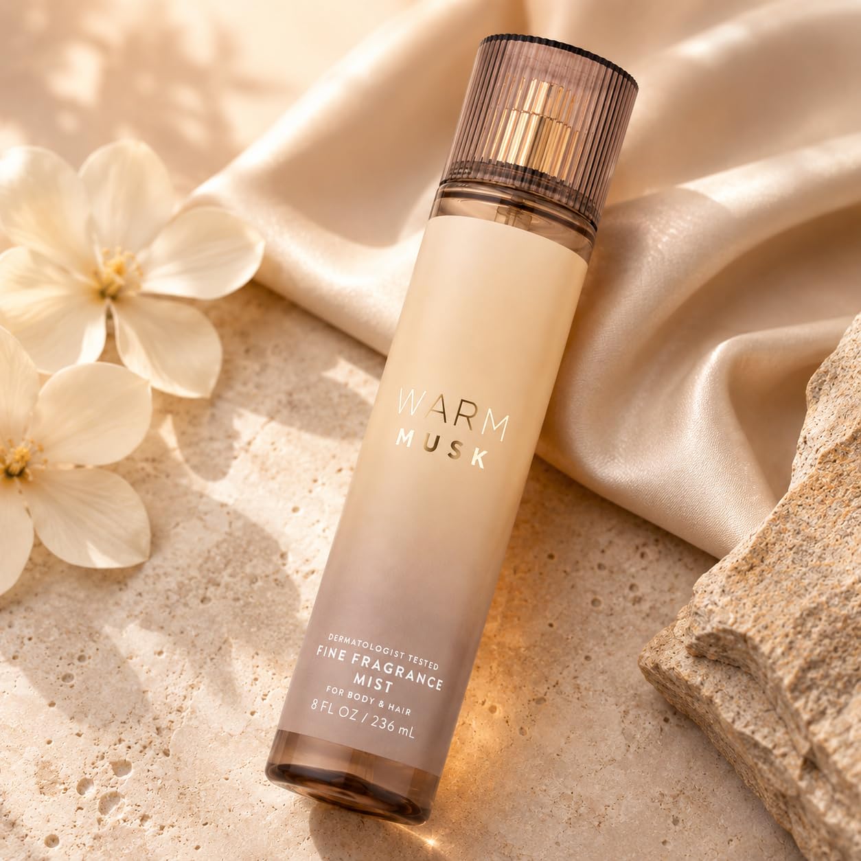 A bottle of Warm Musk Fine Fragrance Mist by Fragrance Tree, styled on a textured stone surface with draped satin fabric and white flowers.