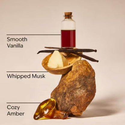 A styled fragrance ingredient composition on a warm beige background, showing a small glass bottle with deep red liquid, vanilla beans, a pat of whipped cream, a large brown rock, and a glossy amber droplet. Text labels read 'Smooth Vanilla,' 'Whipped Musk,' and 'Cozy Amber.