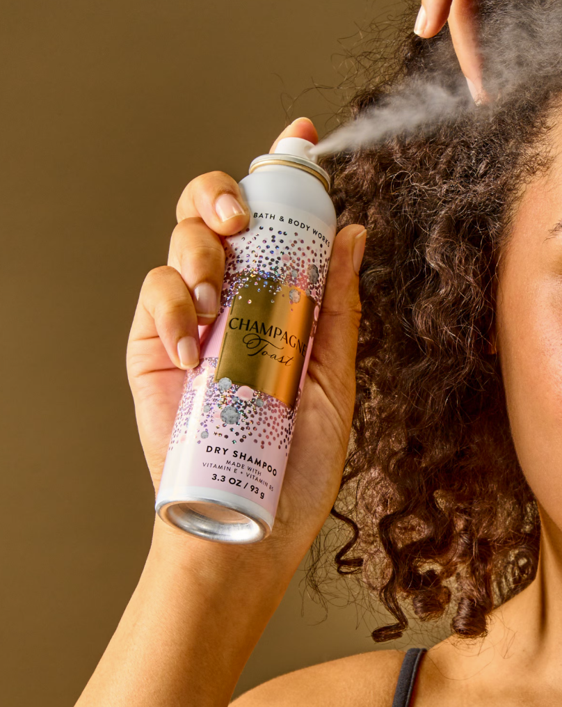 A Pakistani girl sprays Bath & Body Works Champagne Fjord Dry Shampoo onto curly brown hair, with the white and gold aerosol can clearly visible against a warm brown background.