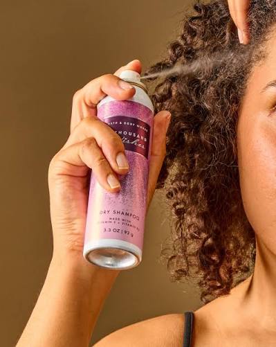 A Pakistani girl with curly dark hair sprays bath and body works dry shampoo from a pink and white aerosol can against a warm brown background.