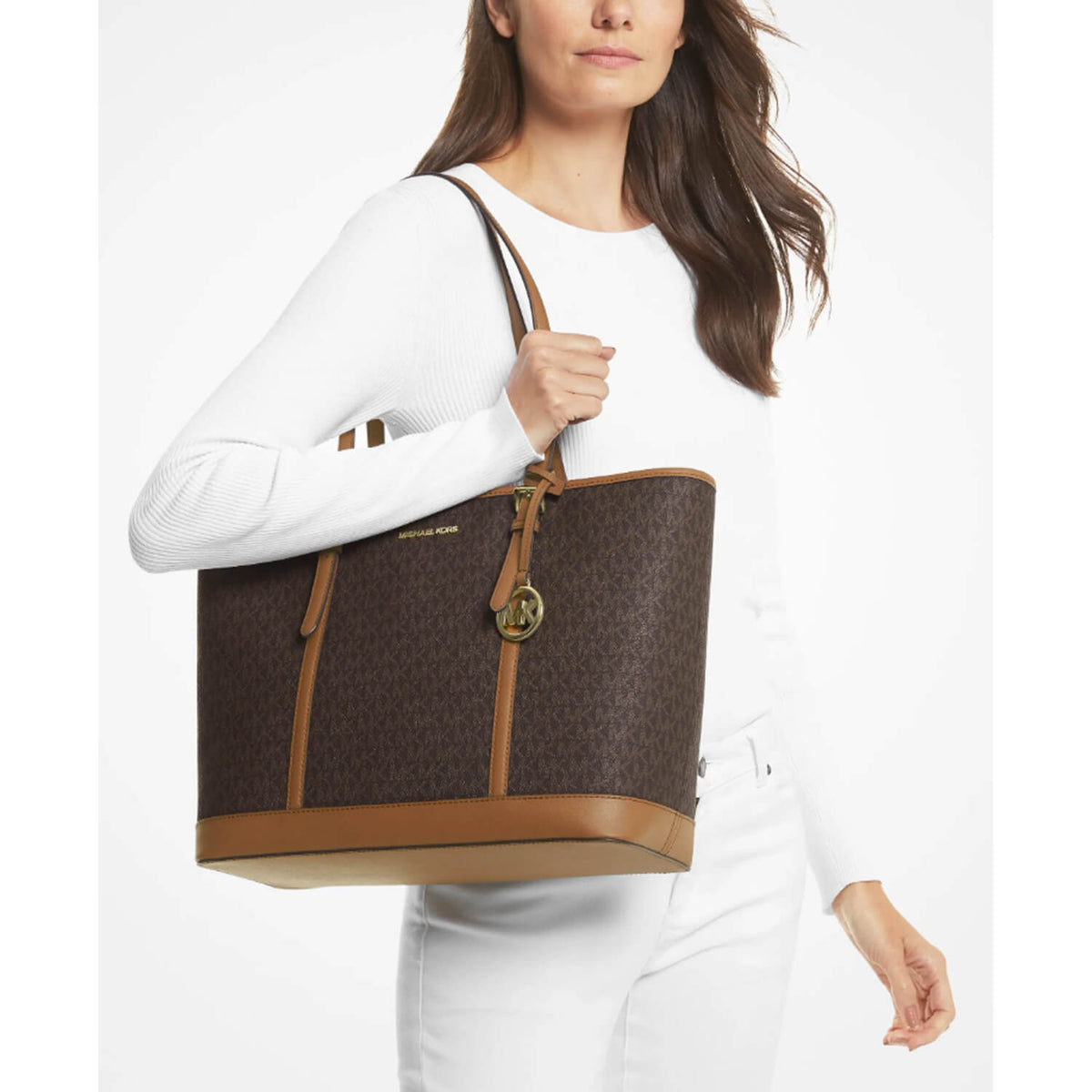 A model carries a Michael Kors tote brown bag. The bag features a dark brown monogram pattern with tan leather trim, a top handle, and a gold-tone circular logo charm.