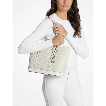 A model carries a light beige Michael Kors monogram tote bag over her shoulder against a plain white studio background.